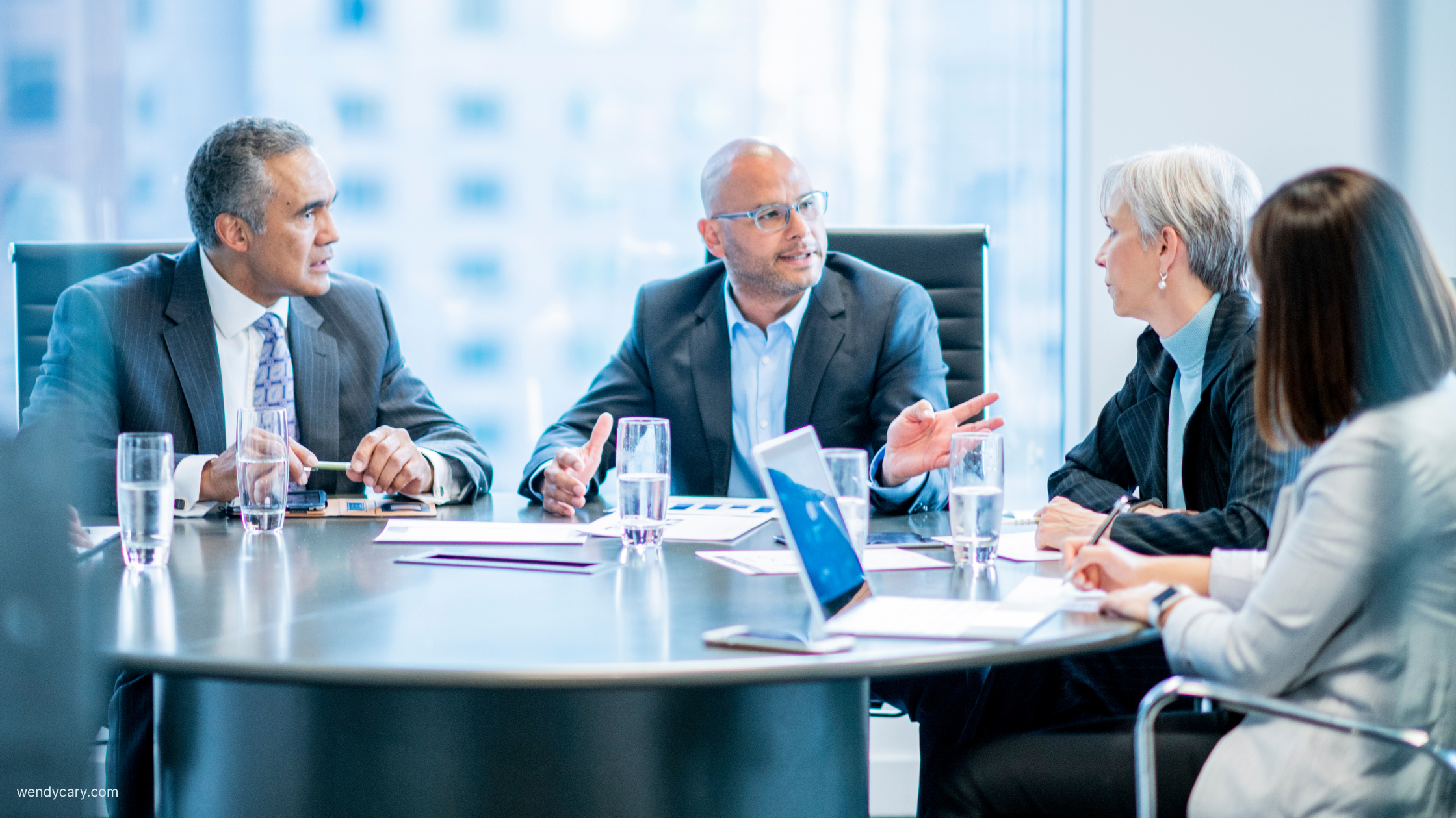 Four senior professionals in a conference room discussion, with one man speaking as the others listen.